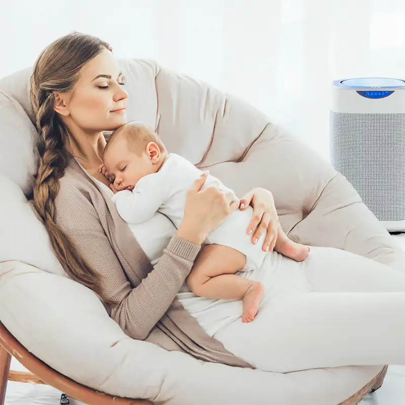 Mother cradling sleeping baby in cozy chair with air purifier improving indoor air quality — Lincoln Heating & Air, Springfield, IL